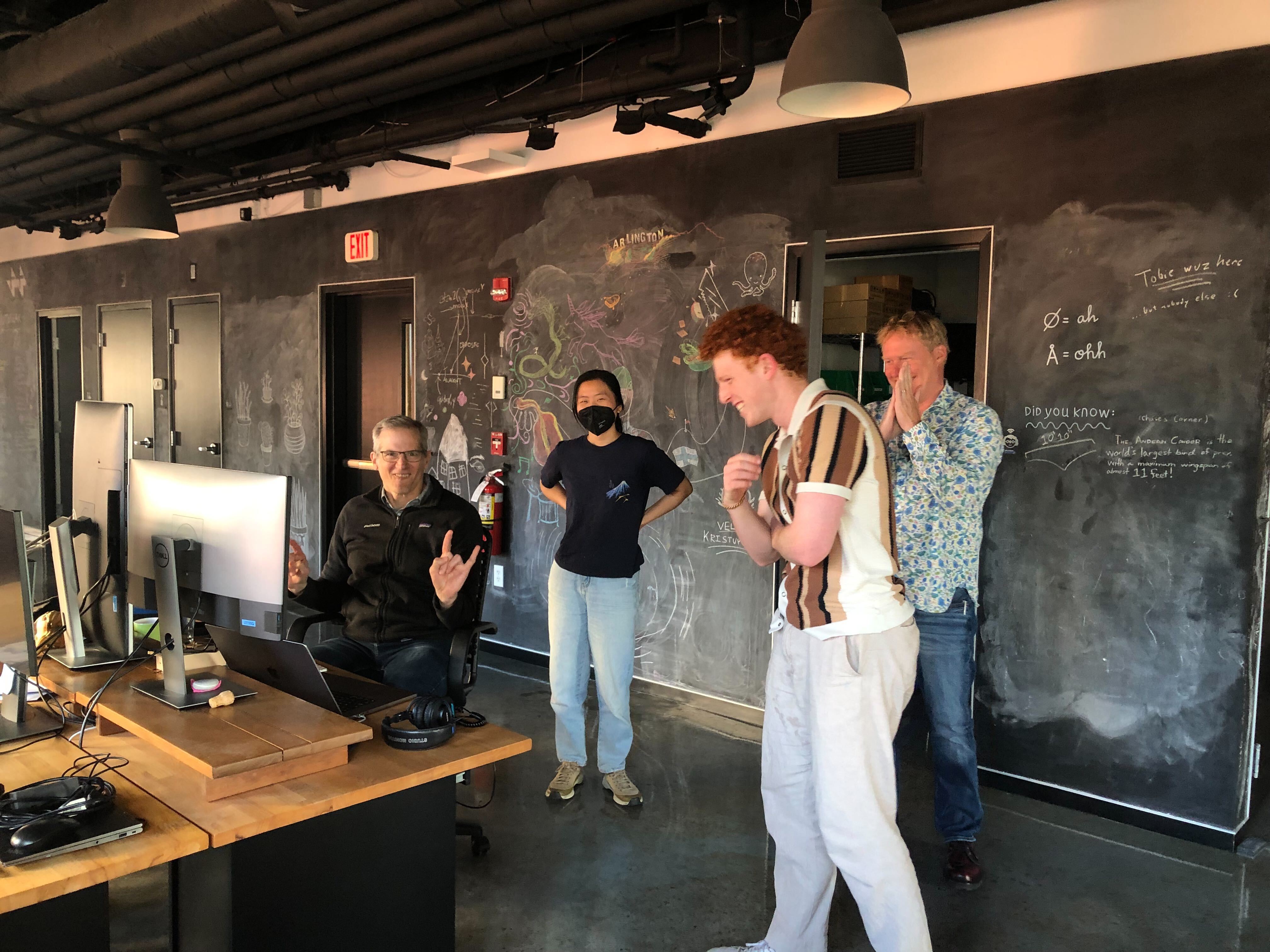 Mark making a rockstar hand gesture while seated as his desk, with Katherine, Ben, and Andy standing around him and smiling.